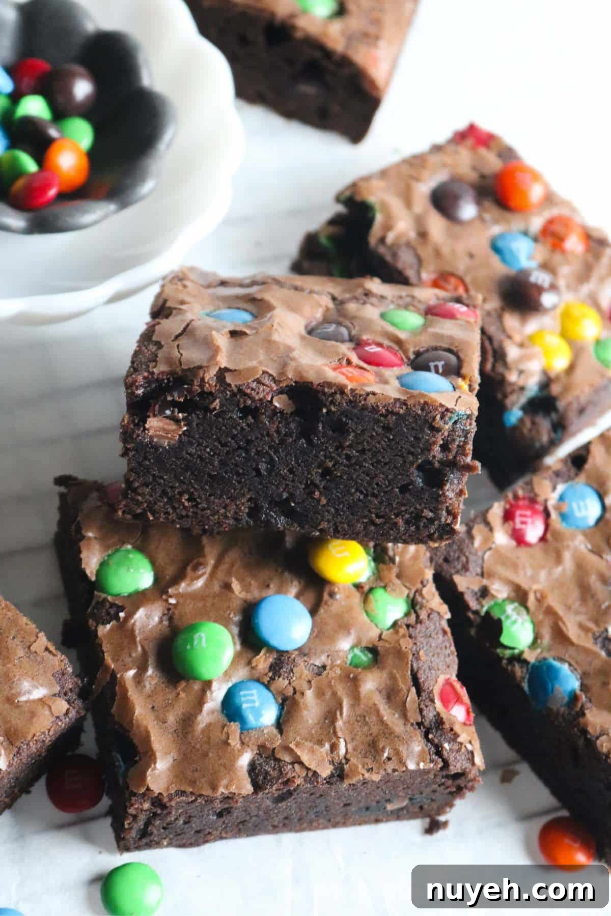 stack of homemade m&ms brownies shingled together on a white surface with a bowl of candies in the background.