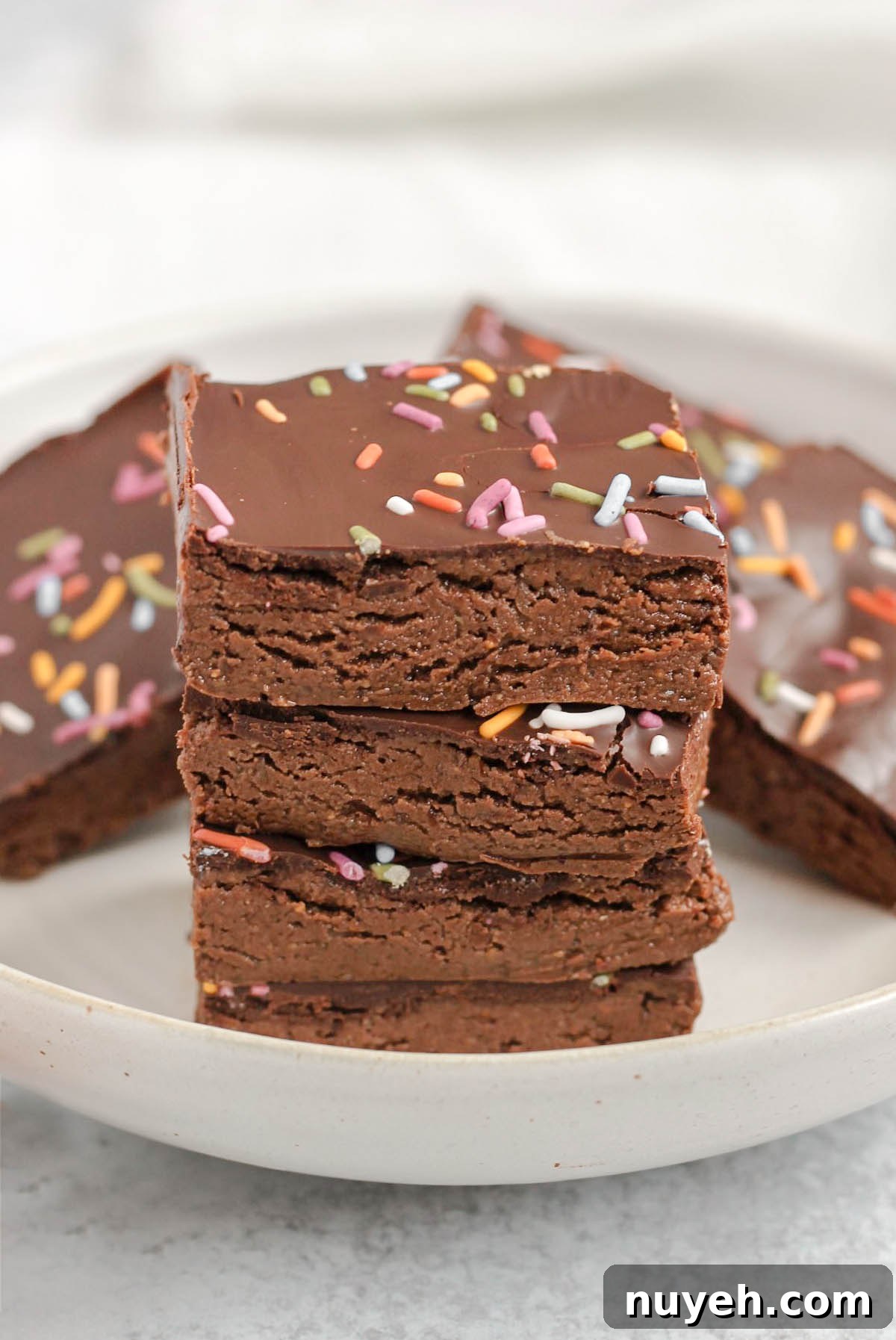 Square pieces of chocolate protein bar stacked 4 high on a plate with other bars in the background.