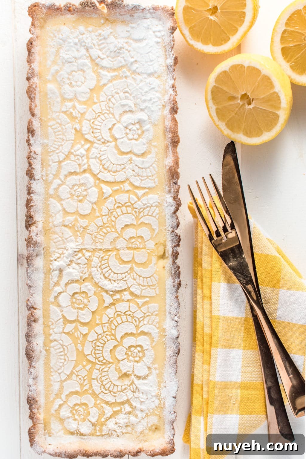 A top down photo of a rectangular lemon tart, with a gingham yellow napkin and fresh lemons.