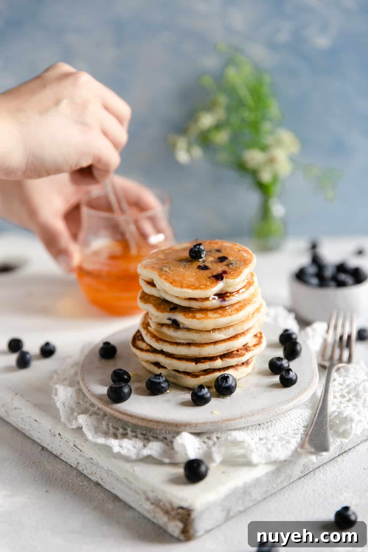 Straight ahead shot of small stack of blueberry pancakes with someone holding glass jar with agave nectar in the background