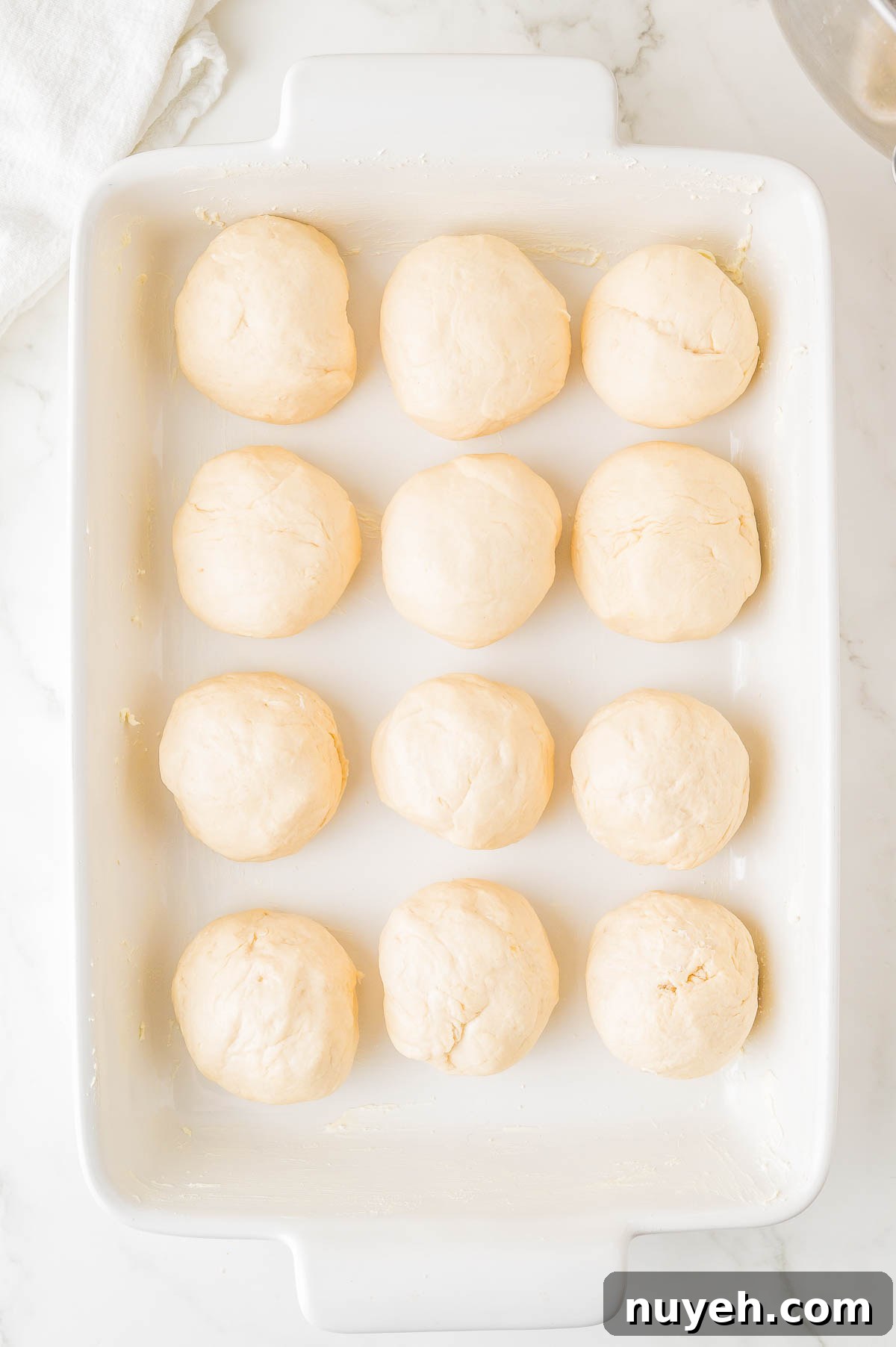 Twelve raw dough balls arranged in rows in a rectangular white baking dish.