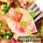 A close-up of Bavarian potato salad with bacon pieces, red onions, green onions, and garnished with fresh dill, being lifted by a metal fork.