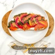 Toasted bread spread with cream cheese and topped with herb-cured salmon, pickled red onion slices, preserved lemon slices and pickled mustard seeds on a white plate with a butcher block and white marble background.