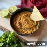 A bowl of pineapple habanero salsa sits on a rustic wooden cutting board surrounded by limes, cilantro, and a red linen.