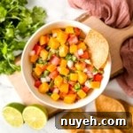 A white bowl of mango pico de gallo surrounded by tortilla chips, limes, cilantro, and a pink linen.