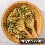 A wooden bowl filled with orzo pesto salad mixed with arugula, sun-dried tomatoes, feta cheese, and herbs, accompanied by two wooden serving utensils.