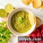 A bowl of serrano salsa is shown with limes, chips, cilantro, and a red linen.