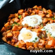 A cast iron skillet containing a sweet potato hash with diced sweet potatoes, pieces of bacon, fresh herbs, and two sunny-side-up eggs on top.
