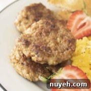 A plate containing three cooked sausage patties, scrambled eggs, sliced strawberries, and a piece of bread.