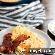 A plate of scrambled eggs, hash browns, and bacon sits on a wooden table, with a striped cloth and a baking dish containing more food in the background. A fork is placed on the plate.