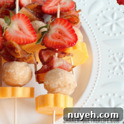 Skewers with strawberries, cheese cubes, bacon pieces, and doughnut holes arranged on a decorative white plate.