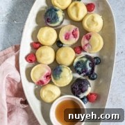 Platter of assorted mini berry cheesecakes and fresh berries with a small bowl of syrup on a gray textured surface, accompanied by a pink cloth.