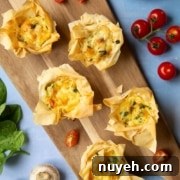 A wooden board with six filo pastry tartlets, each filled with a mixture of vegetables and cheese. Cherry tomatoes and spinach leaves are scattered around, with additional tartlets on a turquoise plate.