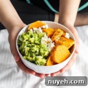 Person holding a bowl containing avocado mash, feta cheese, and sliced sweet potatoes, sitting on a white bedspread.