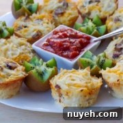 A white plate holds several hash brown and cheese cups, kiwi pieces, and a central square bowl filled with salsa, with a metal spoon resting inside.