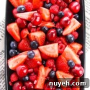 A rectangular dish filled with a variety of fresh red and purple berries, including strawberries, cherries, raspberries, and blueberries, placed on a newspaper.