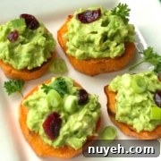 A white plate with four sweet potato slices topped with avocado, chopped green onions, and dried cranberries, garnished with parsley. A fork and green napkin are in the background.