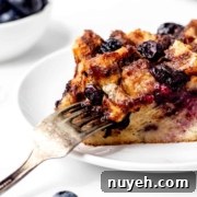 A slice of bread pudding with blueberries on a white plate, with a fork cutting into it. A small bowl of blueberries and a few loose blueberries are in the background.