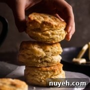 A hand is stacking three golden-brown biscuits on a sheet of parchment paper with another biscuit and butter in the background.