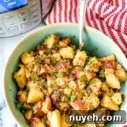 A bowl of cooked potato salad with diced potatoes, herbs, and seasonings sits on a counter next to an Instant Pot. A silver spoon is placed in the bowl.