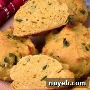 Close-up of several pieces of bread with a golden-yellow color and green herbs visible in the dough, placed on a wooden surface next to a red and white checkered cloth.
