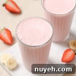Two glasses of pink strawberry banana smoothie on a white surface, with sliced strawberries and bananas around.