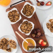 A wooden tray with four oatmeal muffins in paper cups, alongside a honey dipper in a bowl of honey, and a plate with fruit including grapes, strawberries, and pieces of a muffin.