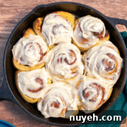 A cast iron skillet filled with eight frosted cinnamon rolls sits on a wooden surface, with a green cloth and one visible cinnamon stick nearby.