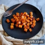 Cubed, sautéed sweet potatoes sit on a black plate with a gold fork on a white marble countertop with an off white linen.