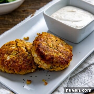 Two cooked salmon patties on a white serving tray with a square bowl of lemon dill sauce.