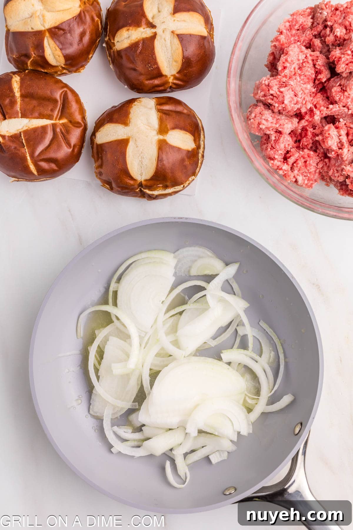 Cooking sliced onions in a skillet in oil with a bowl of ground beef and two pretzel buns
