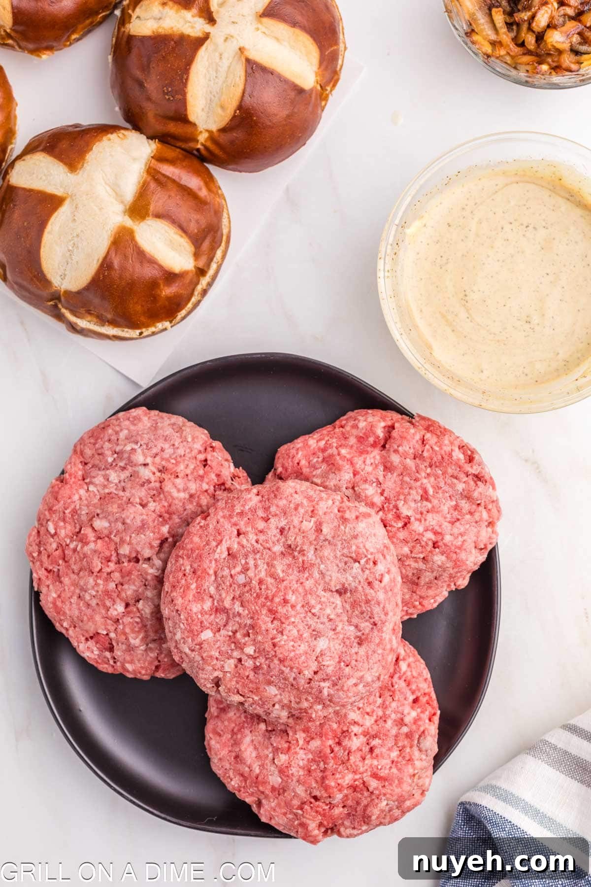 Ground beef patties stacked on a plate with a bowl of sauce and pretzel buns