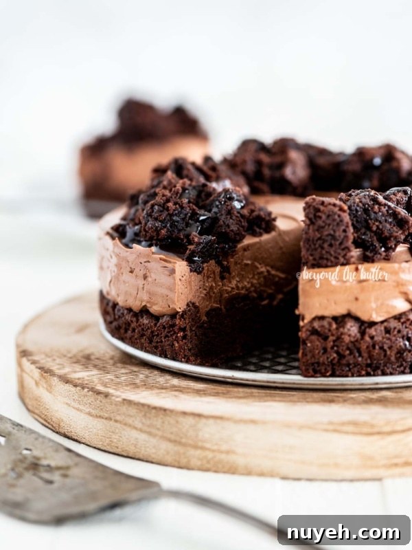 Chocolate brownie cheesecake with slice cut on wood cake plate and cake server in foreground and slice of cake in background on white wood table.