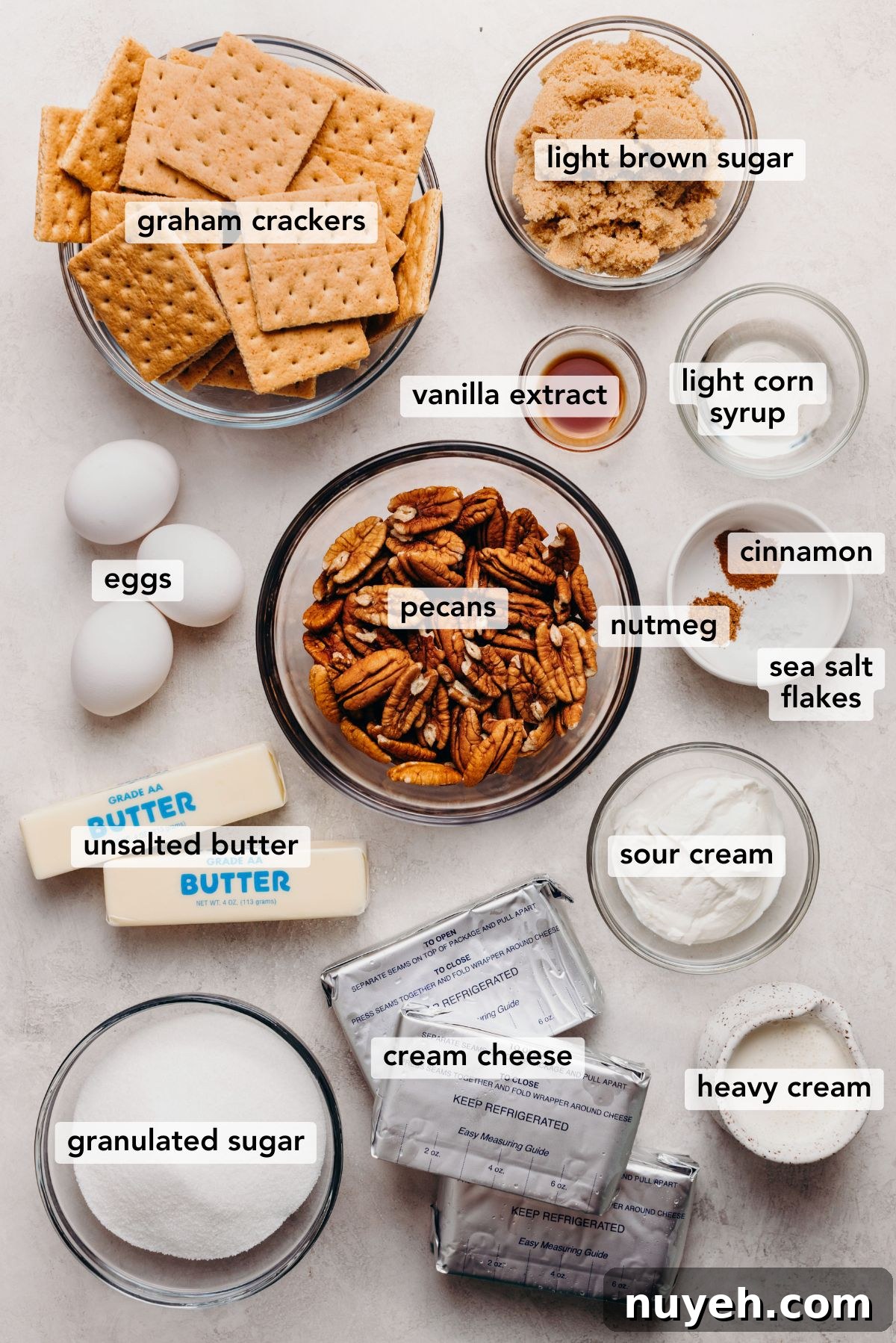 Ingredients in various size bowls and plates labeled as graham crackers, light brown sugar, vanilla extract, light corn syrup, eggs, pecans, nutmeg, cinnamon, sea salt flakes, unsalted butter, sour cream, heavy cream, granulated sugar, and cream cheese on a white marbled background.