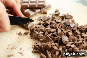 Milk chocolate being chopped up with a black knife on a wooden chopping board.