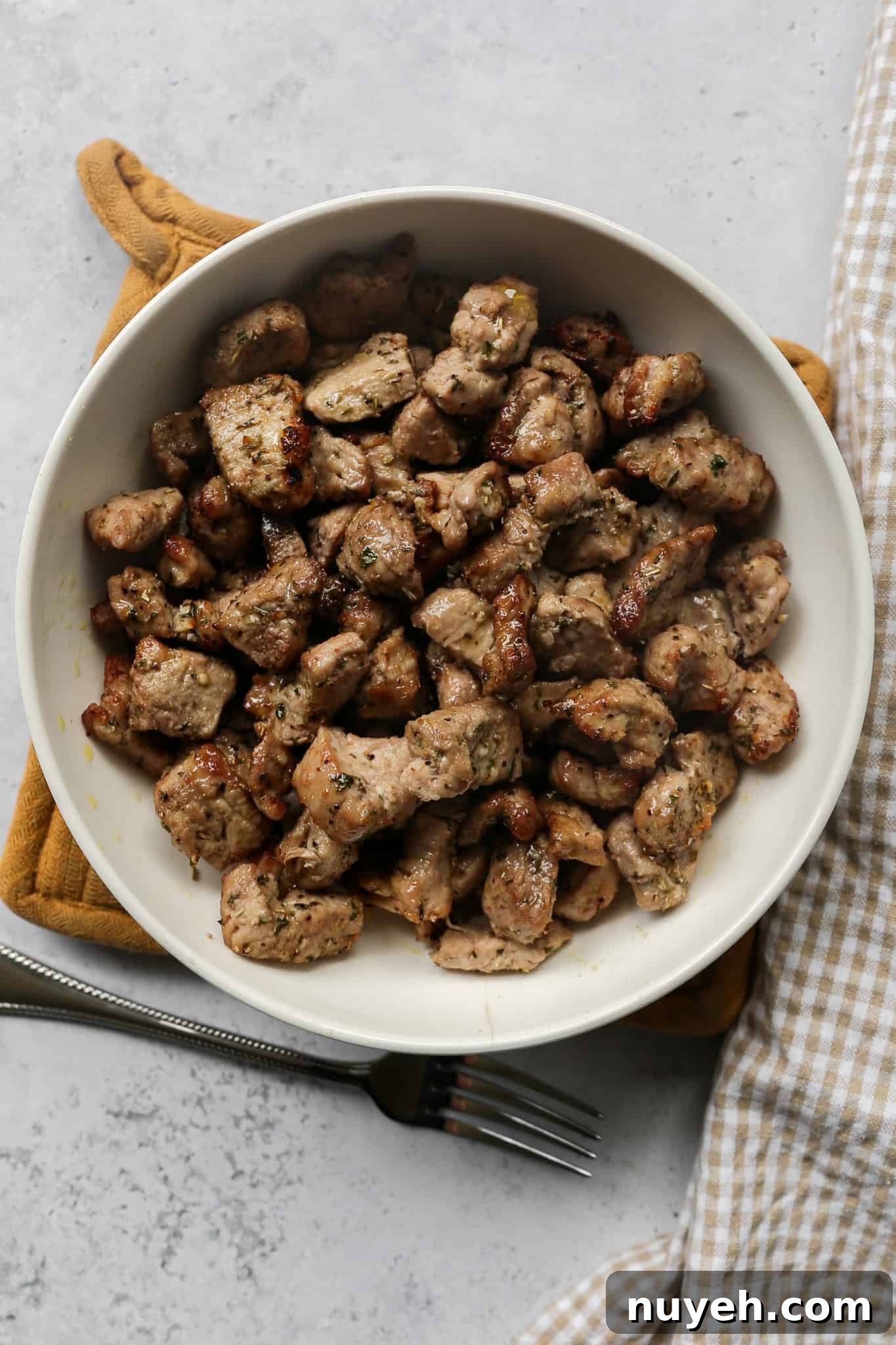 a cream colored serving bowl of air fryer pork tenderloin bites on a tan pot holder next to a silver fork and a tan gingham kitchen towel