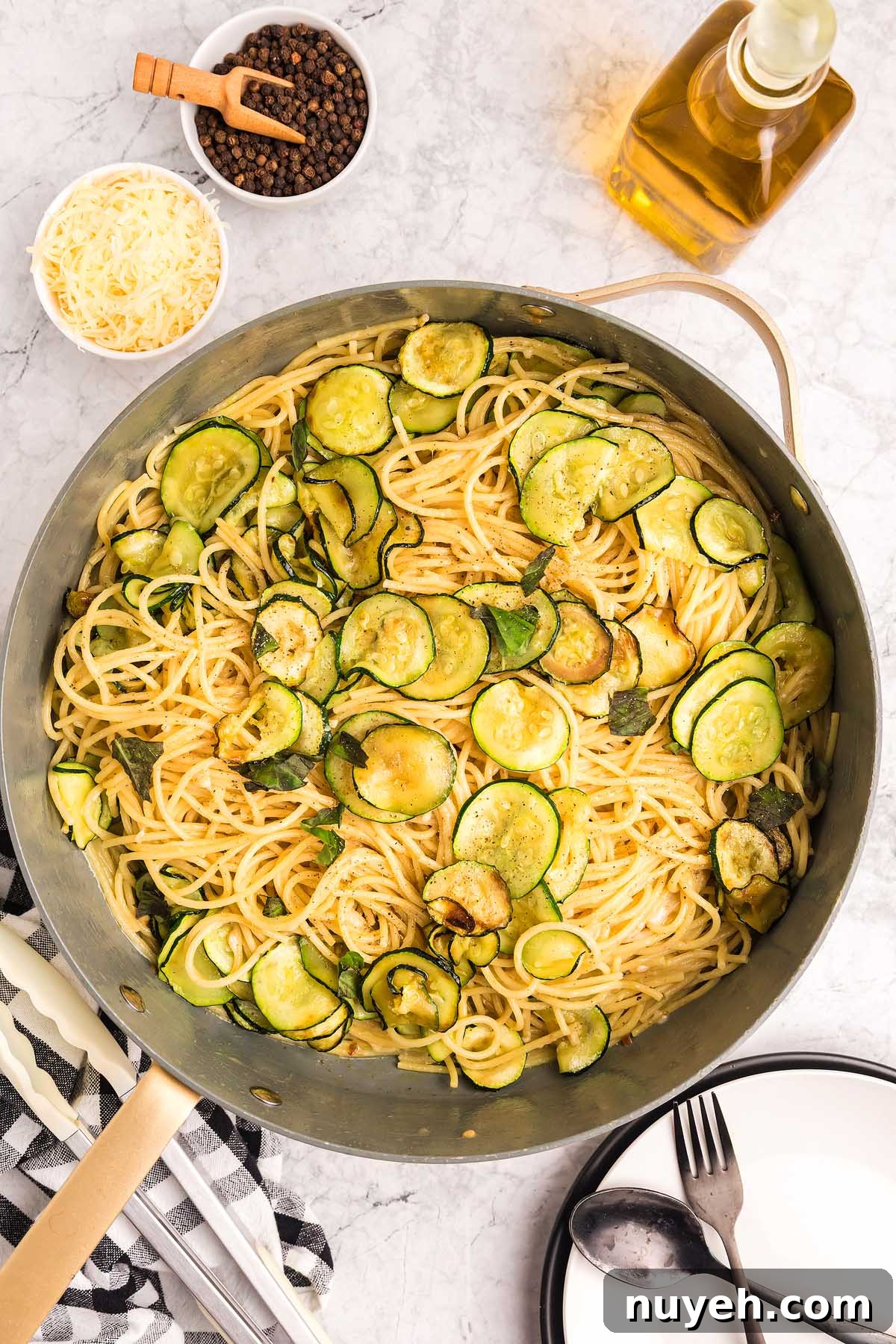 A pan of pasta alla Nerano with sautéed zucchini slices, surrounded by grated cheese, pepper, and olive oil on a marble surface.