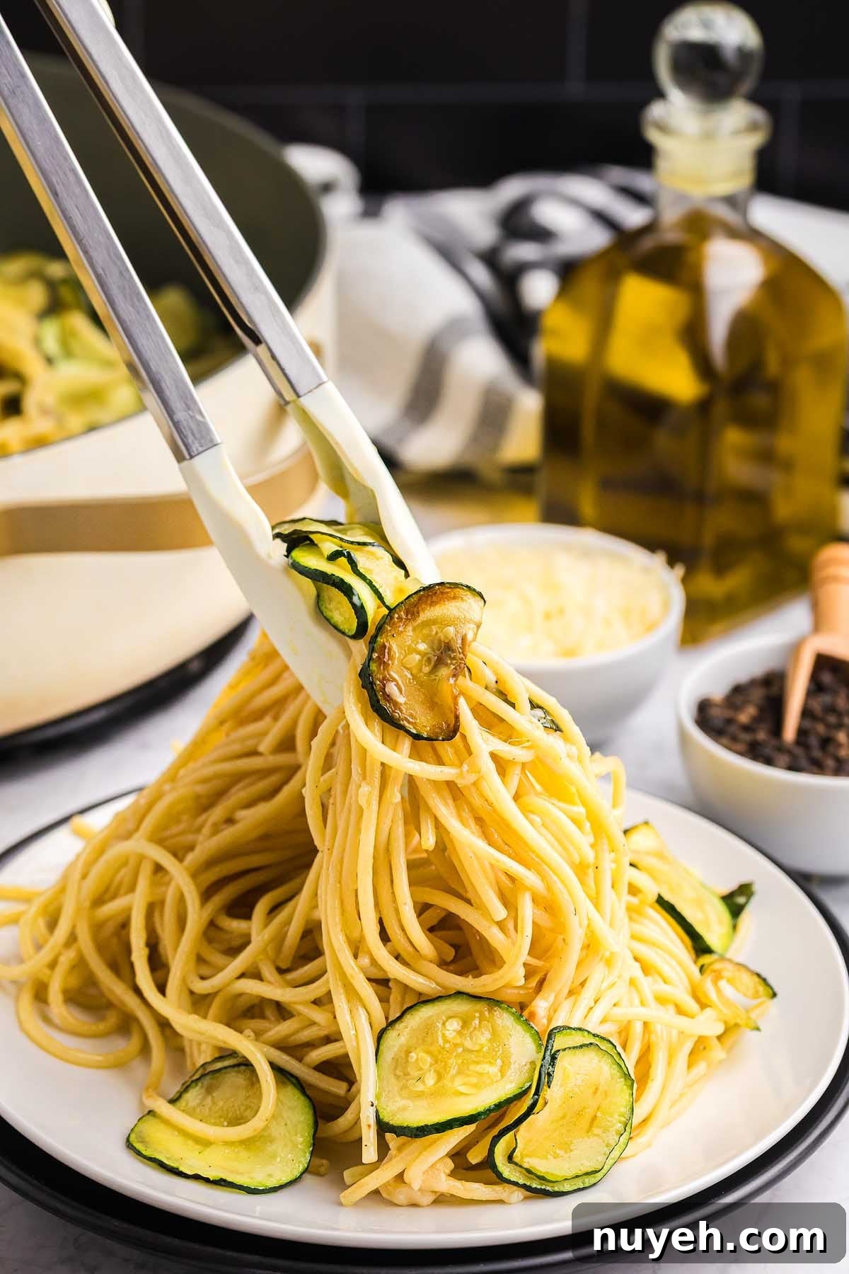 Tongs serving pasta alla Nerano with zucchini slices on a white plate, with olive oil and cheese in the background.