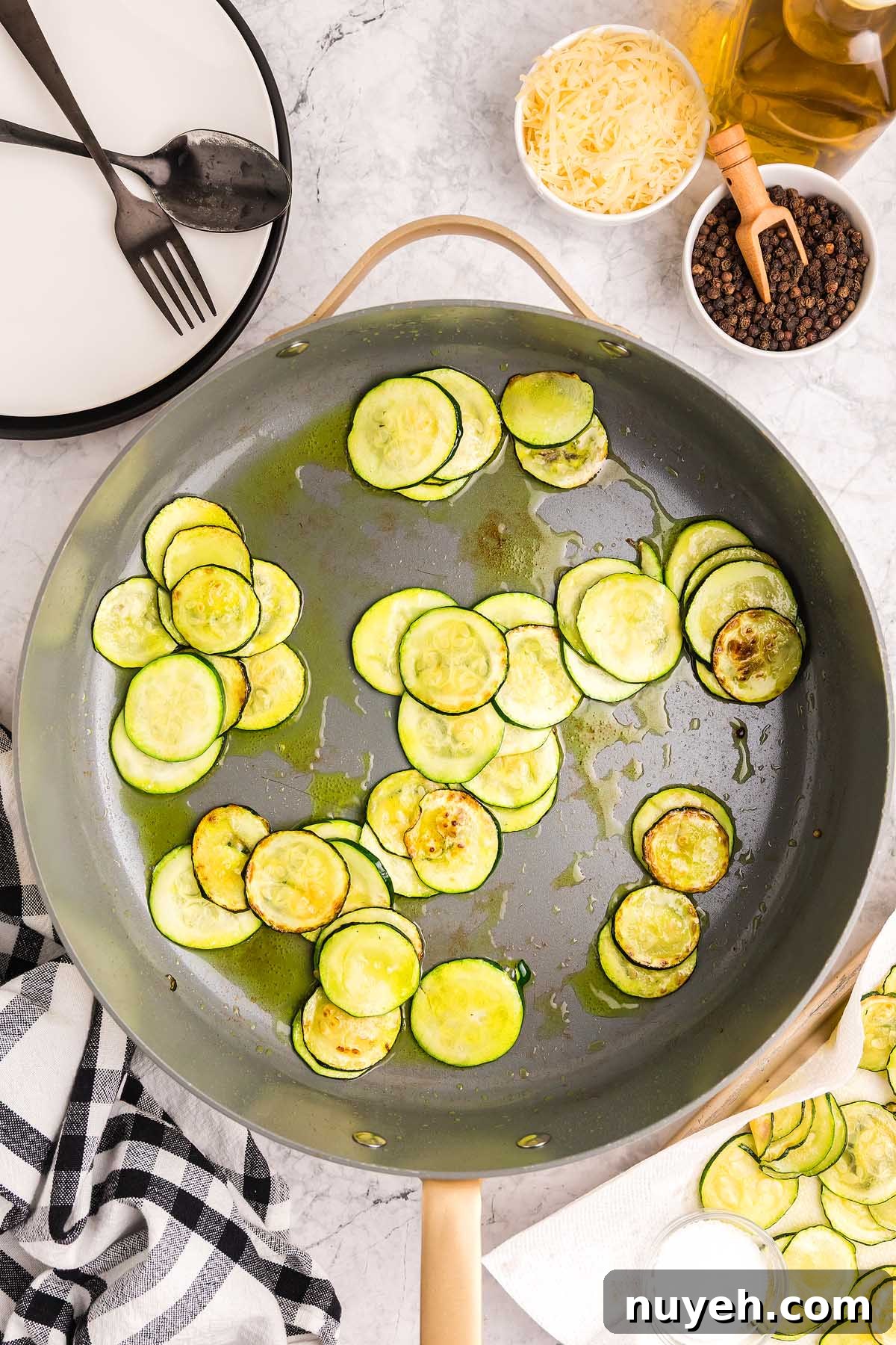 Sliced zucchini sautéing in a skillet, inspired by pasta alla Nerano, with cheese, pepper, and utensils nearby on a marble surface.