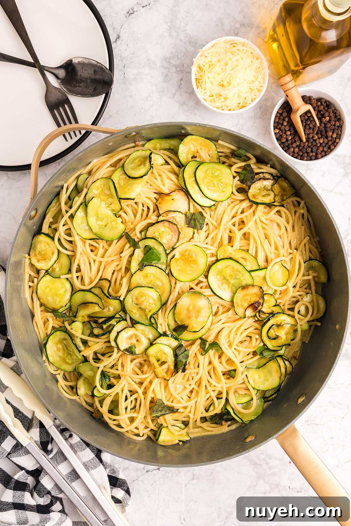 Pasta alla Nerano with sautéed zucchini in a large pan, surrounded by cheese, black pepper, oil, and utensils on a marble surface.