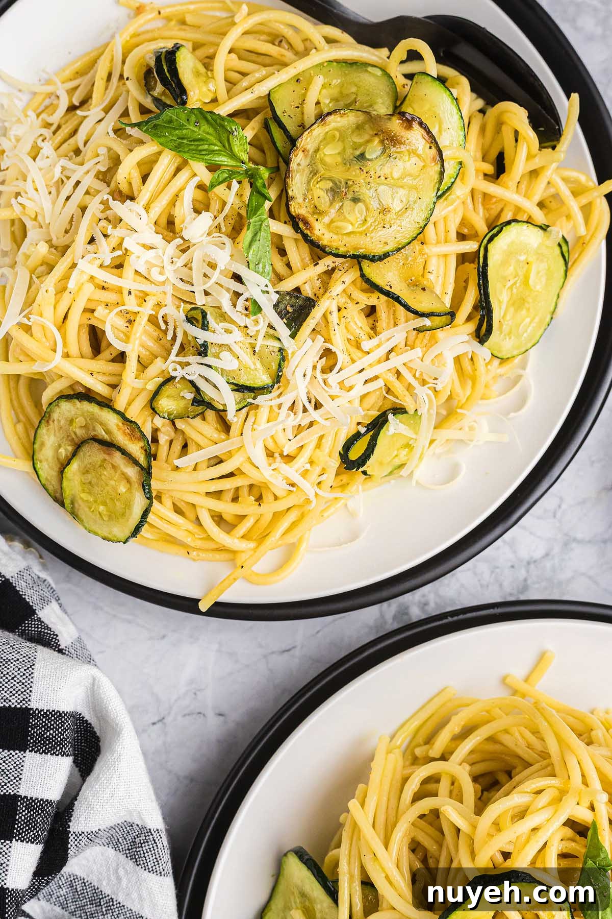 Spaghetti pasta alla Nerano with sautéed zucchini and shredded cheese, served on white plates with a black-and-white napkin.