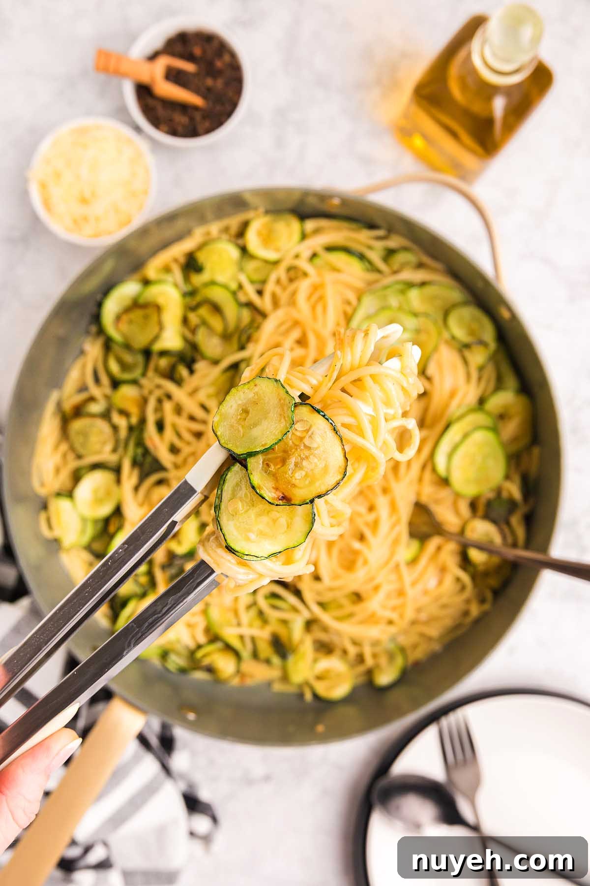 Tongs lifting pasta alla Nerano with sautéed zucchini from a pan, surrounded by ingredients and plates.