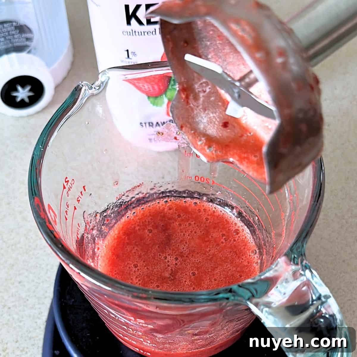 Blending warm strawberries with sugar in a measuring cup with an immersion blender.
