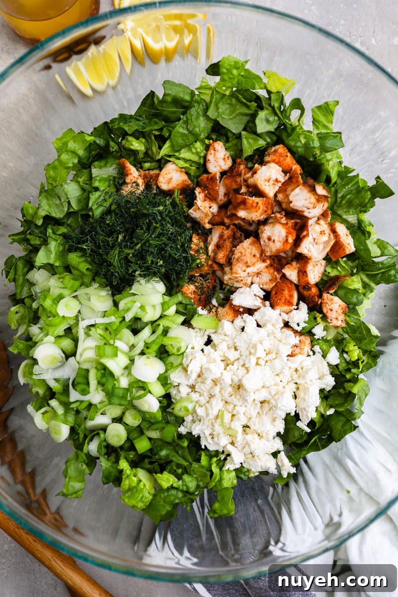 Crisp Chopped Chicken Salad with Feta and Lemon Vinaigrette 5 A large glass mixing bowl filled with chopped romaine lettuce, diced chicken, feta cheese, green onion and fresh dill sitting next to lemon wedges and a jar of lemon vinaigrette