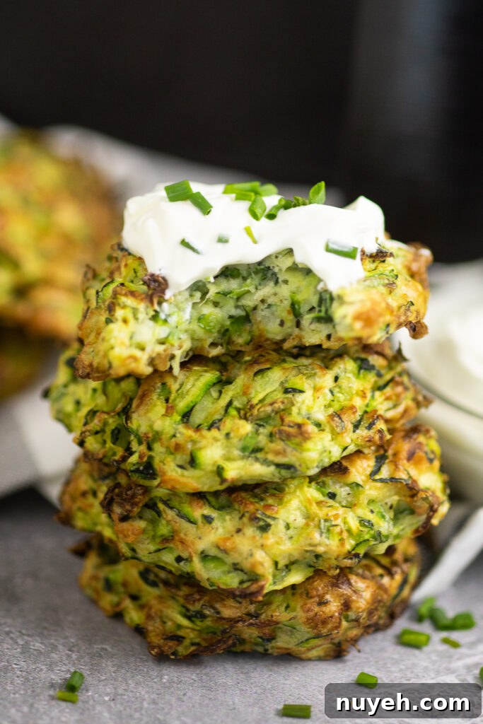 Crispy Air Fryer Zucchini Fritters 2 Stack of 4 air fryer zucchini fritters on a concrete background with a white linen. The top fritter is topped with sour cream and chives and there's a bite taken out