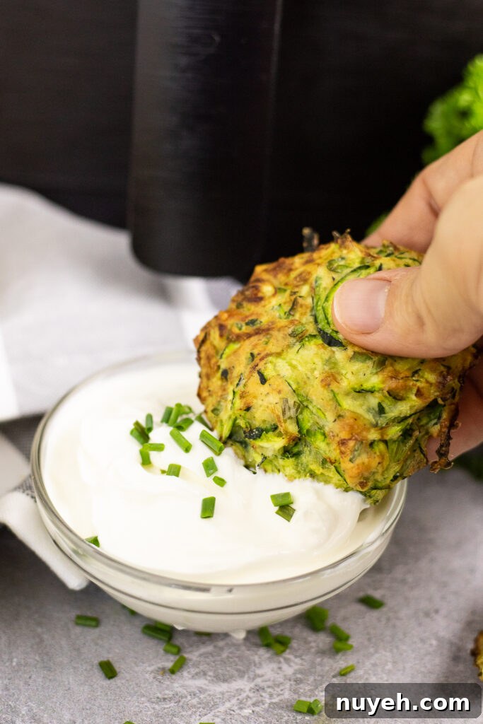 Crispy Air Fryer Zucchini Fritters 3 Air fryer zucchini fritter in front of an air fryer being dipped into a small bowl of sour cream topped with chives.