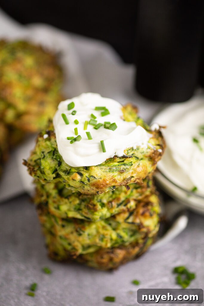 Crispy Air Fryer Zucchini Fritters 4 Stack of 4 zucchini fritters on a concrete backdrop with chives scattered all around. The top fritter is topped with sour cream and chopped chives.