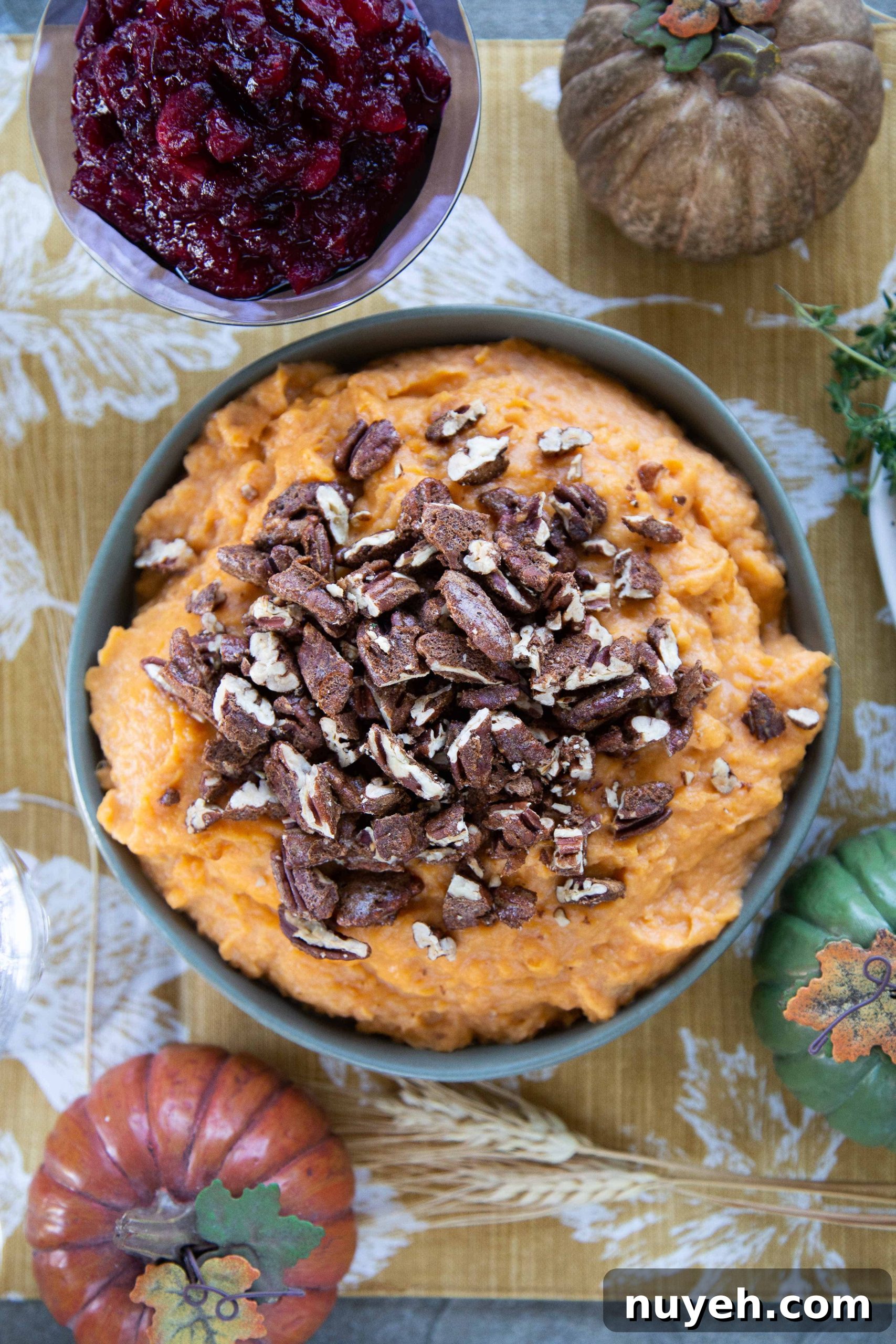 Mashed Sweet Potatoes with Spiced Pecans in a green bowl on a table with cranberry sauce and a gold pumpkin.