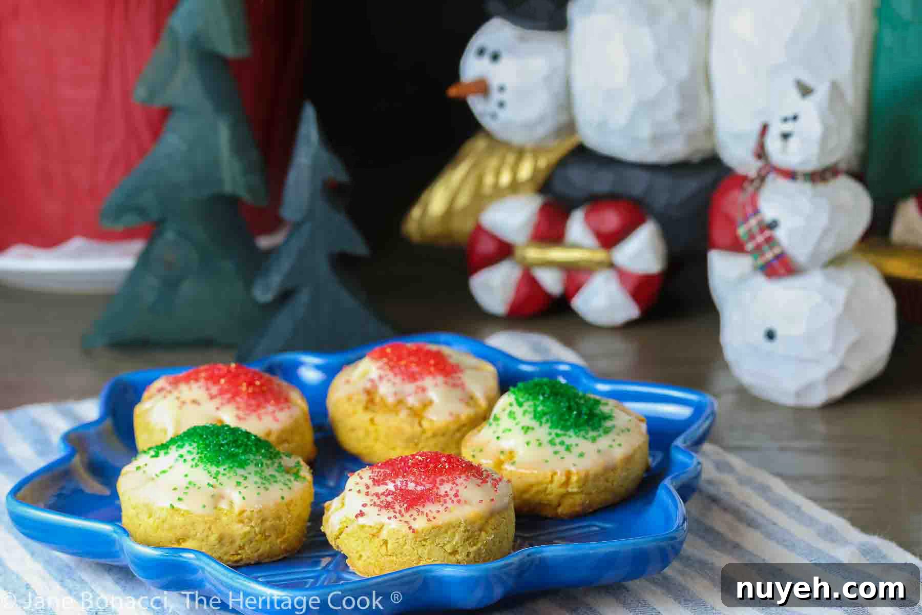 Five White Chocolate Sugared Scones with white chocolate glaze and sprinkled with red and green sugars, served on a blue star-shaped plate on top of a blue and white striped cloth, in front of festive hand-carved snowmen figures © 2023 Jane Bonacci, The Heritage Cook.