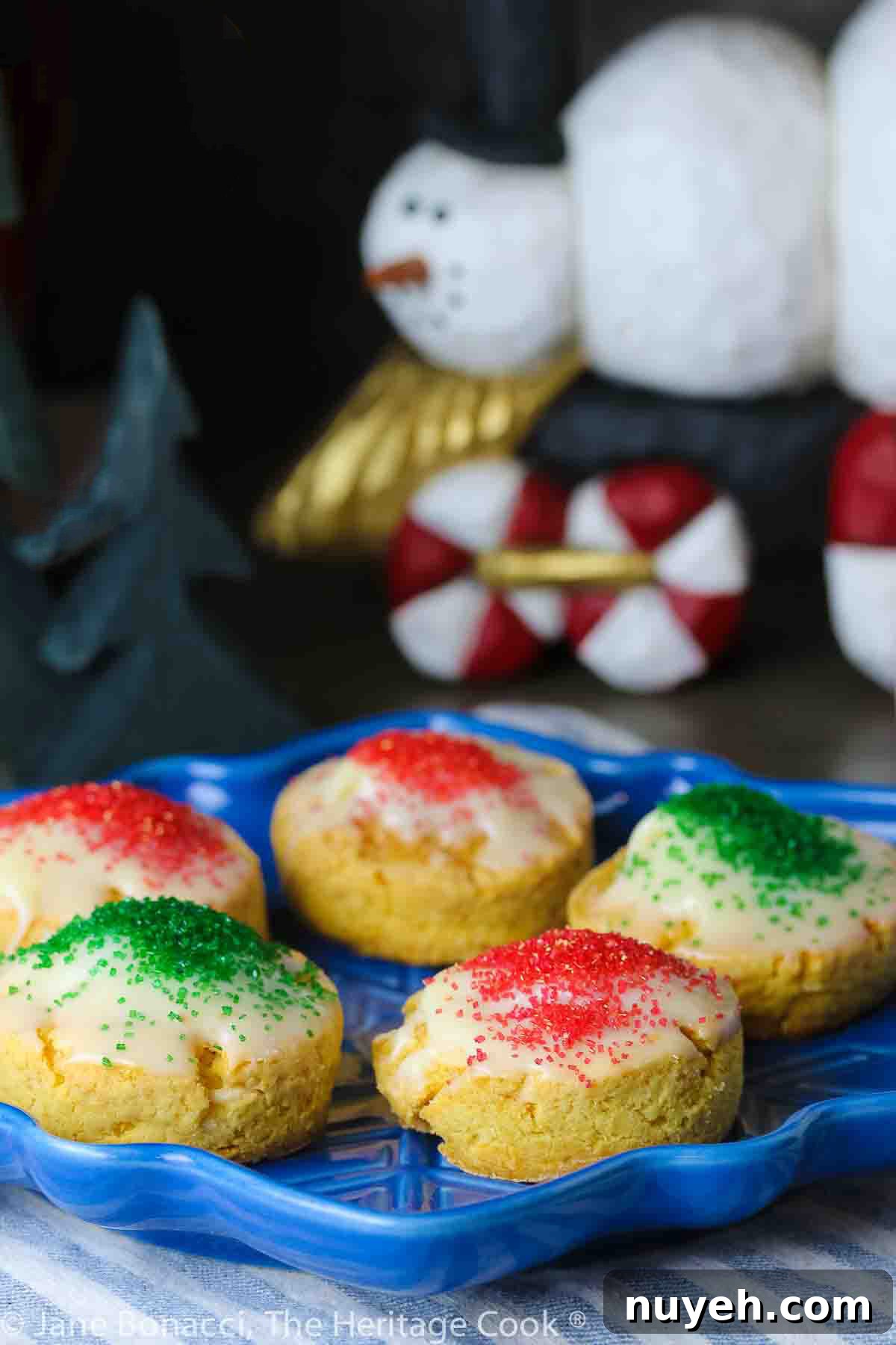 Five White Chocolate Sugared Scones with white chocolate glaze and sprinkled with red and green sugars, served on a blue star-shaped plate on top of a blue and white striped cloth, in front of festive hand-carved snowmen figures © 2023 Jane Bonacci, The Heritage Cook.